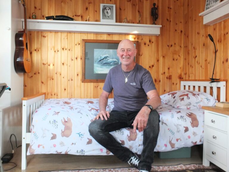 Smiling older man sitting on a bed in a pine-panelled room with a guitar hanging on the wall