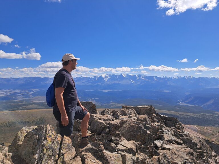 A hiker standing on a rocky summit in the Altai looking towards the snow-capped North Chuysky ridge