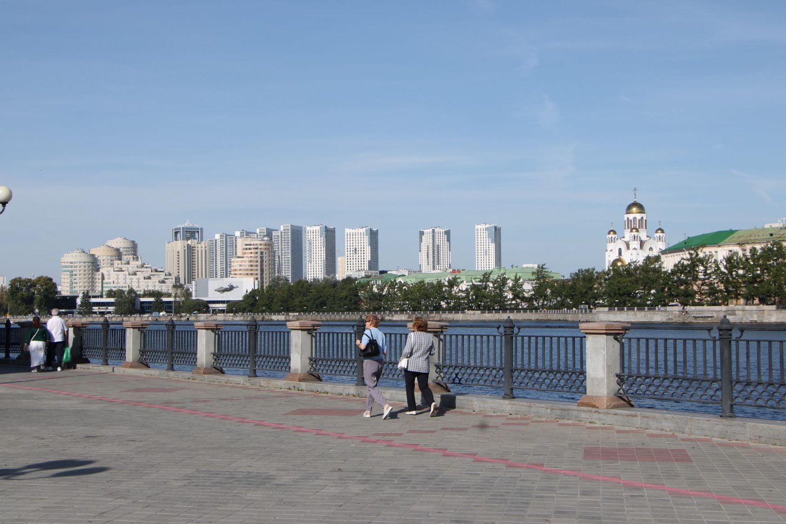 People walking along the river embankment in Yekaterinburg, with tower blocks and a church in the background